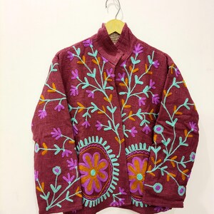 Könnte beinhalten: Eine kastanienbraune Jacke mit Stehkragen und langen Ärmeln, verziert mit gestickten Blumenmustern in Hellblau, Lila und Orange. Die Stickerei umfasst verzweigte Stängel und große kreisförmige Blumenmotive auf den Vorderteilen.