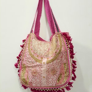 Può includere: Una borsa a spalla rosa con intricati ricami dorati, dettagli in paillettes e un'abbottonatura centrale. La borsa è decorata con nappe rosa e piccole perline bianche lungo i bordi, completa di due lunghi manici in tessuto di colore rosa.