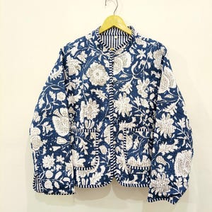 Peut inclure: Une veste matelassée avec un motif floral bleu foncé et blanc imprimé au bloc. La veste présente un col officier, des manches longues, deux poches plaquées à l'avant et une fermeture boutonnée. Les bords sont finis par un liseré rayé bleu et blanc.