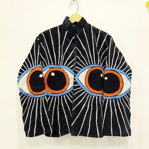 Könnte beinhalten: Eine schwarze Langarmjacke mit einem großen gestickten Augenmotiv auf der Vorderseite. Die Augen haben orangefarbene Iris, schwarze Pupillen und hellblaue Umrandungen, mit weißen gestickten Strahlen, die sich über den schwarzen Stoff erstrecken.