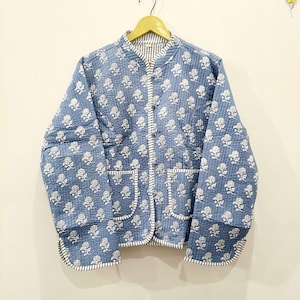 Puede incluir: Una chaqueta acolchada de color azul polvoriento con un patrón floral blanco repetido. La prenda cuenta con cuello mao, mangas largas, dos bolsillos delanteros de parche y un cierre de botones con ribetes a rayas en azul y blanco.