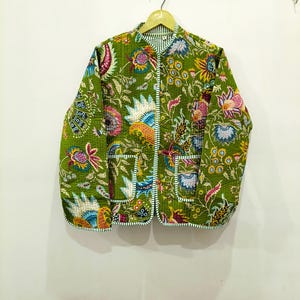 Op de afbeelding: Een doorgestikt jack met lange mouwen in een levendige olijfgroene basiskleur, voorzien van een veelkleurige bloemen- en paisleyprint. Het jack heeft een ronde halslijn, een knoopsluiting aan de voorzijde en twee opgestikte zakken met witte strepen.