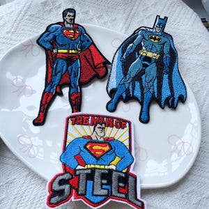 May include: Three embroidered patches featuring comic book characters. One patch shows Superman in a blue suit with a red cape, another shows Batman in a blue and grey suit with a cape, and the third is a shield-shaped patch with Superman and the text THE MAN OF STEEL.