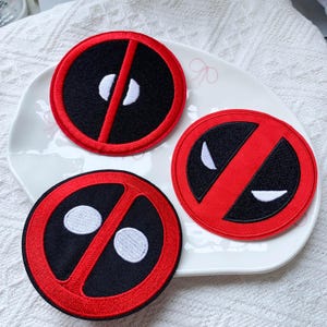 May include: Three circular embroidered patches featuring a red and black design. Two patches display white circular eyes, while one patch features white angular, narrowed eyes. Each patch has a thick red border and a central red diagonal stripe.