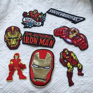 May include: A collection of eight embroidered patches featuring Iron Man imagery, including a Stark Industries logo, a Stay Gold banner, a skull-style mask, and various character poses in red and gold thread with black outlines.