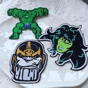 May include: Three embroidered fabric patches featuring comic book characters. One patch depicts a green muscular figure in blue shorts, another shows a green-skinned character with dark hair, and the third displays a helmeted figure in gold and blue.