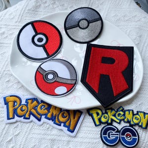 May include: A collection of embroidered patches featuring two circular red and white Poké Balls, one circular grey Poké Ball, a black shield-shaped patch with a red letter R, and two yellow and blue Pokémon logo patches, one with the text Pokémon GO.