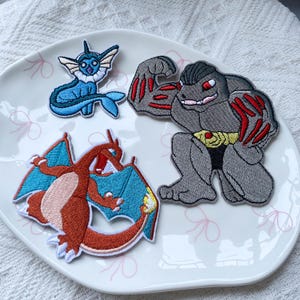 May include: Three embroidered fabric patches featuring cartoon creatures. One patch depicts a small blue and white aquatic creature, another shows a large orange dragon with blue wings, and the third displays a muscular grey humanoid figure with red accents.