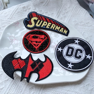 May include: Four embroidered fabric patches featuring DC Comics themes. One patch displays the word SUPERMAN in yellow, one is a circular red and black Superman logo, one is a circular black and white DC logo, and one is a red and black bat shape.