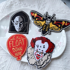 May include: Four embroidered patches featuring horror-themed designs: a black and white vampire portrait, a moth with yellow and red wings, a red balloon patch with the text "WE ALL FLOAT DOWN HERE," and a clown face with red hair and white makeup.