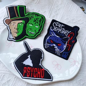 May include: Three embroidered patches featuring horror movie themes. One patch shows a split-face character in a top hat, another is a square patch with a cat and the text PET SEMATARY, and the third is a black silhouette with the red text PSYCHO.