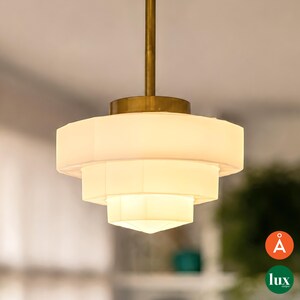 Può includere: Una lampada a sospensione a gradini con tre paralumi in vetro bianco di forma esagonale impilati. L'apparecchio è sospeso da un'asta centrale in metallo color ottone e include un rosone circolare abbinato in cima ai livelli di vetro.