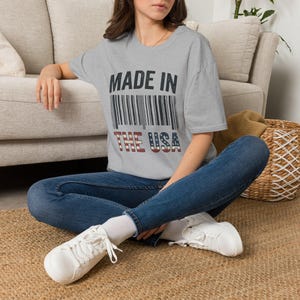 Peut inclure: Un t-shirt gris chiné à manches courtes avec le texte noir MADE IN au-dessus d'un code-barres et le texte THE USA dans un motif de drapeau rouge, blanc et bleu. Le t-shirt est porté avec un jean bleu foncé et des baskets blanches à lacets.