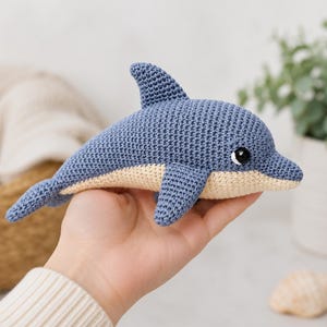 May include: A small, handmade crochet dolphin toy featuring a textured blue upper body and a light beige underbelly. The dolphin has a prominent dorsal fin, side fins, a tail, and a single black and white plastic safety eye visible on the side.