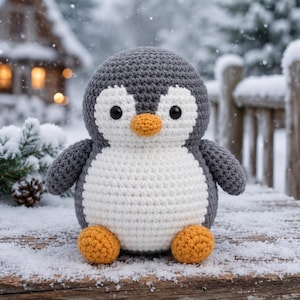 May include: A round, crocheted penguin plush toy featuring a dark grey head and back, a white belly and face patch, two small black eyes, and a bright orange beak and feet. The stuffed animal has a soft, textured yarn construction throughout.