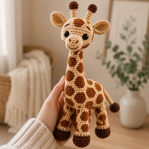 May include: A crochet giraffe toy featuring a light tan body with dark brown spots, two small ossicones on top of the head, large black safety eyes, and dark brown hooves on all four legs. The giraffe has a long neck and a small tail with a tuft.