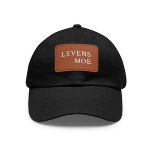 Op de afbeelding: Zwarte baseballpet met een gebogen klep. Een bruin rechthoekig label op de voorkant toont de tekst "LEVENS MOE" in het wit. De pet is gemaakt van een zachte stof.