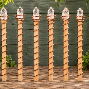 May include: Seven vertical wooden garden stakes arranged in a row, each topped with a clear, pointed quartz crystal point. A copper-coloured metal wire is wrapped in a spiral pattern around the length of each wooden stake from top to bottom.