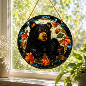 Könnte beinhalten: Ein runder Buntglas-Sonnenfänger mit einem schwarzen Bärenjungen, das zwischen leuchtend orangefarbenen und roten herbstlichen Ahornblättern ruht. Das Motiv ist von einem dunkelblauen und grünen Rand umgeben und hängt an einer goldenen Kordel.