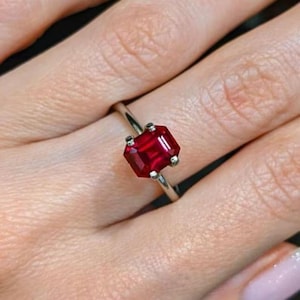 Peut inclure: Une bague argentée ornée d'une pierre précieuse rouge taille émeraude fixée par quatre griffes. La pierre rouge vive est rectangulaire avec des bords facettés, maintenue par des griffes métalliques sur un anneau fin et poli entourant le doigt.