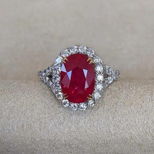May include: A ring featuring a large, oval-cut, deep red ruby centre stone set in yellow gold prongs. The ruby is surrounded by a halo of round, brilliant-cut white diamonds set in a silver-toned metal band, with additional diamonds along the shank.