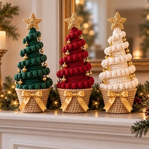 Op de afbeelding: Drie kegelvormige gehaakte kerstbomen in donkergroen, dieprood en wit. Elke boom heeft een gouden ster als piek, kleine gouden bolvormige ornamenten in een spiraal langs de zijkanten en een beige gehaakte voet met een gouden satijnen strik.