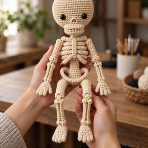 Könnte beinhalten: Eine beige gehäkelte Skelettpuppe mit rundem Kopf, schwarzen Perlenaugen, Brustkorb, beweglichen Armen und Beinen sowie einer Beckenstruktur. Die Figur ist aus Garn gefertigt und weist eine deutlich sichtbare Maschenstruktur auf.
