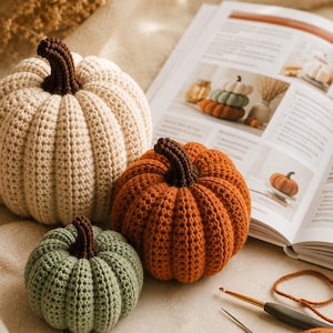 May include: Three crocheted pumpkins in cream, orange, and sage green, each featuring a dark brown stem. The pumpkins are arranged near an open instructional book, a metal crochet hook, a sewing needle, and a ball of orange yarn.