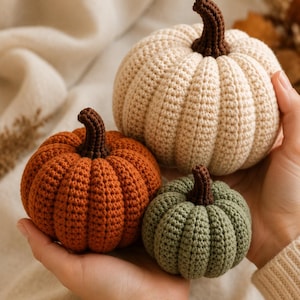 May include: Three crocheted pumpkins in cream, orange, and sage green colors. Each pumpkin features a textured, ribbed surface and a dark brown crocheted stem. The pumpkins are round and vary in size, with the cream one being the largest.