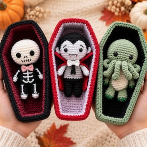 May include: Three crocheted dolls inside individual coffin-shaped containers. The left doll is a white skeleton in a black suit with a pink bow tie. The center doll is a vampire with a red-lined cape. The right doll is a green octopus-like creature.