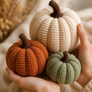 May include: Three crocheted pumpkins in cream, burnt orange, and sage green. Each pumpkin features a textured, ribbed surface and a dark brown crocheted stem. The pumpkins are round and vary in size, showcasing detailed yarn stitching throughout.