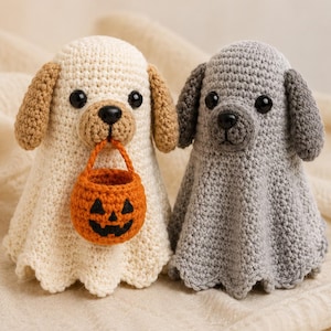 May include: Two crocheted dog figures, one cream-colored and one gray, both shaped like ghosts with rounded heads and flared bodies. The cream dog has tan ears and holds a small, orange, crocheted jack-o'-lantern bucket in its mouth.