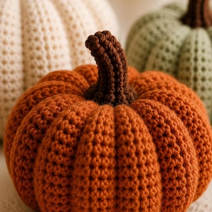 May include: Three crocheted pumpkins in orange, cream, and sage green. Each pumpkin features a textured, ribbed surface and a dark brown crocheted stem. The orange pumpkin is in the foreground, while the cream and green ones are positioned behind.