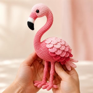 May include: A crocheted flamingo toy featuring a long, curved neck, a black and white beak, and a body covered in layered, scalloped feathers in shades of light pink, medium pink, and dark pink, standing on two thin, textured pink legs.