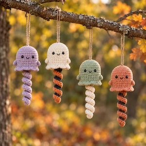 May include: Four crocheted ghost ornaments hanging from a tree branch. Each ghost has a rounded head with black bead eyes and a stitched smile, featuring a long, spiraling tail. The ghosts are colored light purple, cream, sage green, and orange.