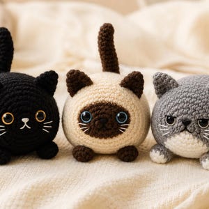 May include: Three round crocheted cat plushies featuring a black cat with amber eyes, a cream and brown Siamese-style cat with blue eyes, and a grey and white cat with a grumpy expression, each with a small upright tail and embroidered whiskers.