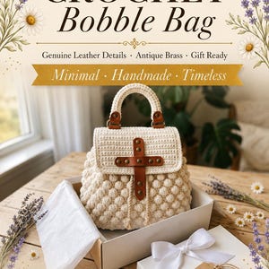 May include: A cream-coloured crochet bobble bag featuring a top handle and a flap closure secured with a brown leather strap and antique brass hardware. The bag is presented in an open box with white tissue paper and a white ribbon bow.