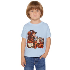 May include: A light blue short-sleeve t-shirt featuring a graphic of a squirrel wearing a witch hat, a ghost, and other small animals surrounded by pumpkins and autumn leaves, with the text "NUT OR TREAT" printed in a bold, stylized font.