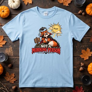 Peut inclure: Un t-shirt bleu clair avec un motif représentant un renard déguisé en vampire tenant un petit parapluie sous un soleil brillant. Le renard est assis près d'un panneau indiquant NO SUN PLEASE. Sous le renard, le texte indique NOT A MORNING PERSON.