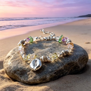 Puede incluir: Una pulsera de plata con un dije de concha y una mezcla de conchas, cuentas transparentes y coloridas. La pulsera descansa sobre una roca gris en una playa arenosa con el océano al fondo.