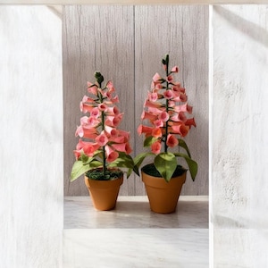 May include: Two artificial foxglove plants in terracotta pots. The pink flowers have a bell-like shape and are arranged on tall stems with green leaves. The pots sit on a white surface against a light wood background.