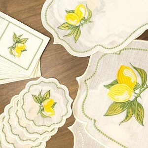 May include: A set of off-white fabric table linens featuring embroidered yellow lemons with green leaves. The collection includes rectangular placemats, a runner, and circular coasters, all finished with scalloped edges and green stitching.