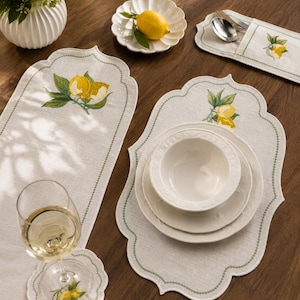 Puede incluir: Un juego de camino de mesa, mantel individual y posavasos de lino beige con bordes festoneados, cenefas bordadas en verde e ilustraciones de limones. Incluye un portacubiertos a juego, un cuenco de cerámica blanca y platos.