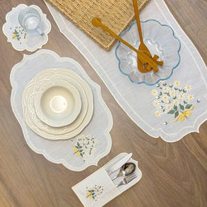 Puede incluir: Un juego de camino de mesa, mantel individual, posavasos y porta cubiertos blanco con bordes festoneados y ramos de margaritas bordados con lazos amarillos. Incluye un cuenco de cristal, cubiertos de madera y platos de cerámica.