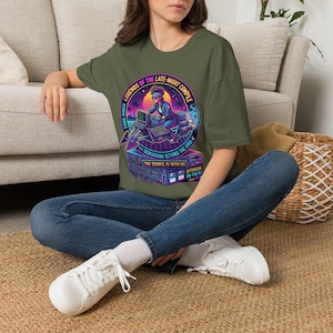 May include: An olive green t-shirt featuring a vibrant graphic print of a person at a computer with the text "LEGENDS OF THE LATE-NIGHT COMPILE," "CODE RIDER," "DEBUGGING BEYOND THE GRID," "THE SOURCE IS WITH US," and "OPTIMIZING ON-THE-FLY."