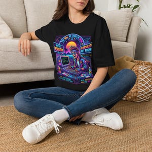 May include: A black t-shirt featuring a vibrant 1980s-style graphic of a person using a vintage computer. The design includes neon colours, palm trees, and text that reads: The 80s: We Coded In Neon, Where Debuggin Is An Absolute Art Form!