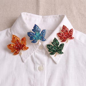 May include: Four maple leaf brooches pinned to a white collared shirt. The brooches are orange, blue, green, and red, each featuring metallic outlines and small embedded crystals that match the color of the enamel leaf surface.