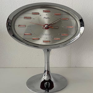 Könnte beinhalten: Verchromter Rhythm-Wecker mit ovalem, silbernem Zifferblatt und roten und weißen Akzenten. Die Zeiger der Uhr sind rot. Die Uhr hat ein modernes Design und steht auf einem Sockel. Hergestellt in Japan.