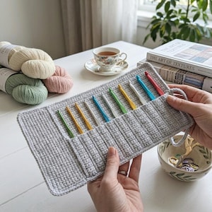 May include: A light gray crocheted organizer case holding a set of ten colorful metal crochet hooks in various sizes. The case features individual vertical slots for each hook, with a matching gray crocheted loop closure on the right side.
