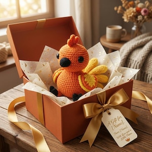May include: A bright orange crocheted phoenix plush toy with black eyes and beak, nestled inside an open orange gift box lined with gold-flecked tissue paper. A gold satin ribbon and a gift tag with the text A Gift for [Name] are attached.